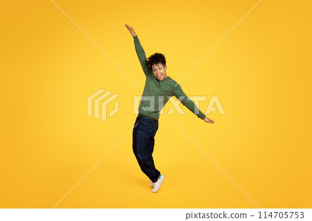 Exuberant young african american man mid-jump with arms outstretched and a joyous expression on yellow background 114705753