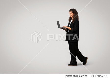 A professional woman in a business suit walks while intently using a laptop, suggesting multitasking and productivity A professional woman in a business suit walks while intently using a laptop, suggesting multitasking and productivity 114705755