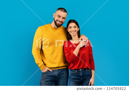 Smiling couple dressed in colorful sweaters standing confidently on blue background, romantic man and woman posing in studio together against bright backdrop, looking at camera, copy space 114705822