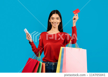 Happy woman holding multiple shopping bags, credit card and smartphone in hands, cheerful female enjoying making purchases, standing on blue background 114705841