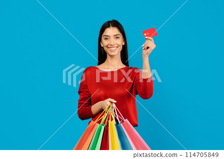 Young happy woman holding credit card in one hand and bright shopper bags in the other, happy female ready for shopping spree, standing on blue studio background, copy space Young happy woman holding credit card in one hand and bright shopper bags in the other, happy female ready for shopping spree, standing on blue studio background, copy space 114705849
