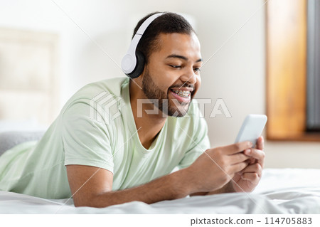 Casual african american man in bed with headphones browsing on his smartphone with focus and enjoyment Casual african american man in bed with headphones browsing on his smartphone with focus and enjoyment 114705883