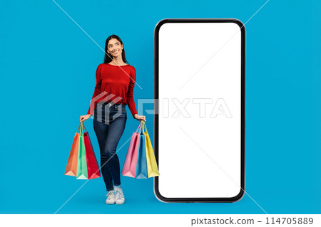 A woman standing with shopping bags in hand near big blank phone, appearing busy and engaged in her shopping activities, mockup A woman standing with shopping bags in hand near big blank phone, appearing busy and engaged in her shopping activities, mockup 114705889
