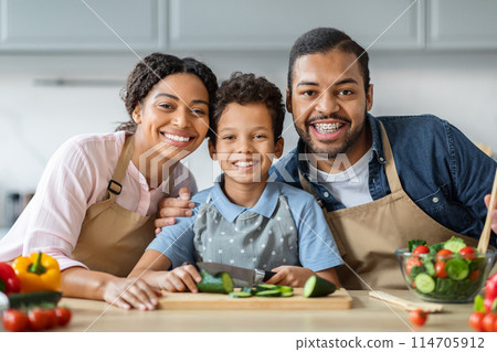 Portrait of cheerful loving african american family cooking together at home. Smiling mother, father and preteen boy kid son preparing healthy meal at cozy modern kitchen, have fun on weekend Portrait of cheerful loving african american family cooking together at home. Smiling mother, father and preteen boy kid son preparing healthy meal at cozy modern kitchen, have fun on weekend 114705912