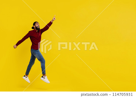 Excited handsome bearded young indian man hipster jumping in the air isolated on yellow studio background, pointing at copy space, showing great online offer or amazing deal, full length 114705931