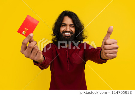Happy long-haired bearded millennial indian guy customer showing red plastic credit card and thumb up, recommending his bank, isolated on yellow studio background. Contactless payment 114705938