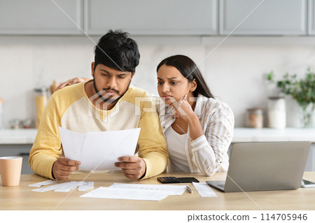 Upset young indian husband and wife sitting at desk in front of computer laptop, reading correspondence, checking bills, millennial family have debts, kitchen interior, copy space 114705946
