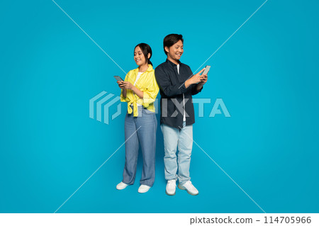 A japanese millennial man and woman stand back to back, each engrossed in their own smartphones, dressed in casual clothing with pleasant expressions against a bright blue background A japanese millennial man and woman stand back to back, each engrossed in their own smartphones, dressed in casual clothing with pleasant expressions against a bright blue background 114705966