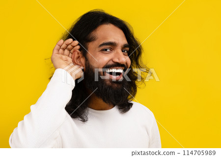Curious interested millennial indian man holding hand near ear and trying to hear information, yellow studio background, copy space. Excited eastern guy in casual clothing eavesdropping 114705989