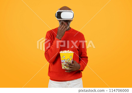 Engrossed senior black man in a red sweater enjoying virtual reality with a headset, snacking on popcorn, and displaying a look of surprise on a yellow background, studio 114706000