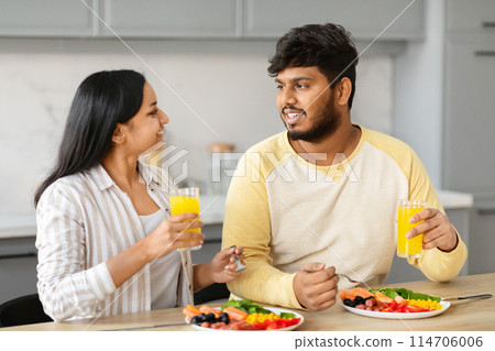Positive young indian spouses enjoying breakfast together at home. Eastern loving couple have meal, drinking fresh orange juice, chatting and smiling, kitchen interior Positive young indian spouses enjoying breakfast together at home. Eastern loving couple have meal, drinking fresh orange juice, chatting and smiling, kitchen interior 114706006
