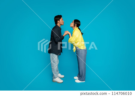 Man in dark jacket and millennial japanese woman in yellow shirt hold hands and lean in close, sharing a moment of playful interaction or conversation on a bright turquoise background 114706010