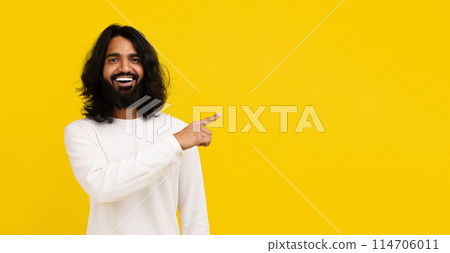 Cheerful bearded long-haired young indian man in white long sleeve pointing at empty copy space mockup for advertisement, isolated on yellow studio background, web-banner for online offer 114706011