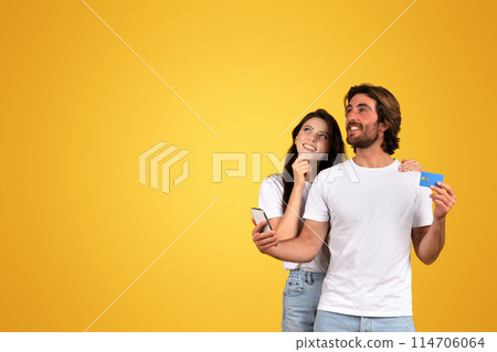 A european woman dreamily looking up and a bearded man smiling while holding a credit card and phone, both in white t-shirts, planning a purchase on a yellow background, studio 114706064