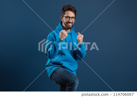 Joyful bearded man in blue hoodie raising fists in triumph, and smiling at camera, happy excited young guy wearing eyeglasses having fun on dark studio background, celebrating success, copy space 114706067