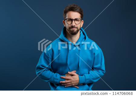 Stomach Pain. Sick young man suffering from acute abdominal ache, upset millennial guy touching belly and frowning, feeling unwell while standing against blue studio background, copy space Stomach Pain. Sick young man suffering from acute abdominal ache, upset millennial guy touching belly and frowning, feeling unwell while standing against blue studio background, copy space 114706068