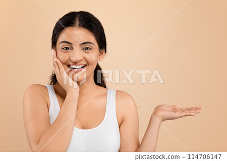 Delighted indian woman in white tank top making presenting gesture, happy young eastern female showing something on open palm and touching face, standing on beige studio background, copy space Delighted indian woman in white tank top making presenting gesture, happy young eastern female showing something on open palm and touching face, standing on beige studio background, copy space 114706147