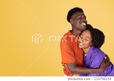 A content smiling black young woman in a purple blouse embraces a joyful young man in an orange shirt, both smiling with closed eyes, sharing a warm moment on a yellow background 114706234