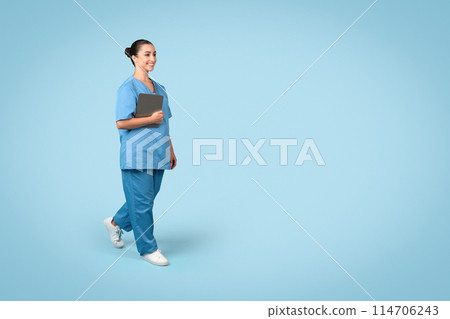 Cheerful young female nurse in scrubs holding tablet while walking, looking positive and professional, blue background, free space, medical banner Cheerful young female nurse in scrubs holding tablet while walking, looking positive and professional, blue background, free space, medical banner 114706243