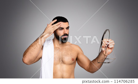 Hot handsome bearded middle eastern young man with towel on his shoulder looking at hand mirror after shower, touching his forehead, grey background. First signs of face skin aging 114706285