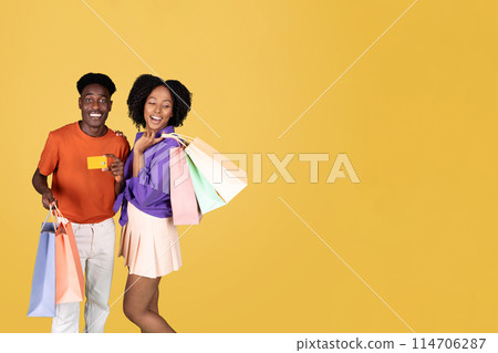 A young couple shopping, the woman in a purple blouse holding colorful bags and the man in an orange shirt showing a credit card, both smiling with satisfaction on a yellow background A young couple shopping, the woman in a purple blouse holding colorful bags and the man in an orange shirt showing a credit card, both smiling with satisfaction on a yellow background 114706287