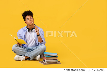 Pensive black male student sits cross-legged with stack of books, holding notebook and pencil, looking at free space, against yellow background, banner Pensive black male student sits cross-legged with stack of books, holding notebook and pencil, looking at free space, against yellow background, banner 114706288