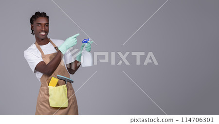 Young handsome black cleaner guy wearing apron pointing aside at copy space, smiling millennial african american man in gloves, advertising cleaning service, standing against grey studio background 114706301