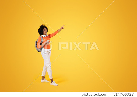 Joyful African American woman with curly hair, pointing with excitement, holding a phone, wearing an orange top, white pants, headphones, and a backpack, on a bright yellow background Joyful African American woman with curly hair, pointing with excitement, holding a phone, wearing an orange top, white pants, headphones, and a backpack, on a bright yellow background 114706328