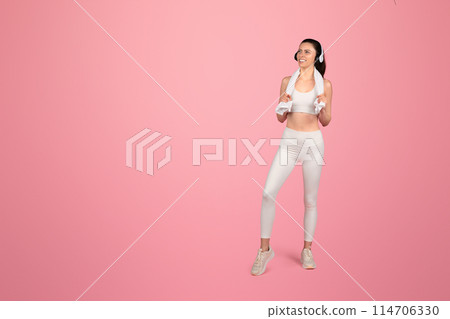 Smiling european fit woman in white sportswear with headphones around neck and towel over shoulders standing confidently on a pink background, radiating health and happiness Smiling european fit woman in white sportswear with headphones around neck and towel over shoulders standing confidently on a pink background, radiating health and happiness 114706330
