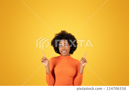 Optimistic African American woman looking up and pointing upwards, expressing positivity, wearing a snug orange turtleneck and white pants, set against a monochromatic yellow background Optimistic African American woman looking up and pointing upwards, expressing positivity, wearing a snug orange turtleneck and white pants, set against a monochromatic yellow background 114706338
