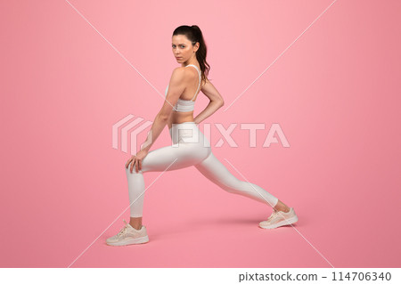 Focused and toned european young woman in a white sports bra and leggings performing a stretching exercise, showcasing strength and flexibility on a pink background, studio 114706340