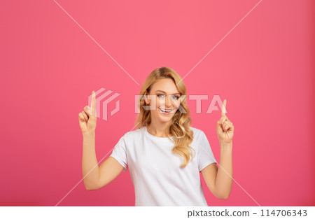 Happy young blonde woman in white t-shirt pointing fingers upwards with both hands on pink studio background. Millennial lady showing her recommendation above head. Free space 114706343