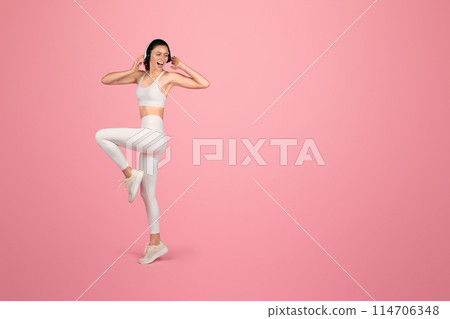 Vibrant and joyful caucasian woman in white sportswear dancing or doing aerobics with headphones, expressing freedom and happiness during a fun workout on a pink background Vibrant and joyful caucasian woman in white sportswear dancing or doing aerobics with headphones, expressing freedom and happiness during a fun workout on a pink background 114706348