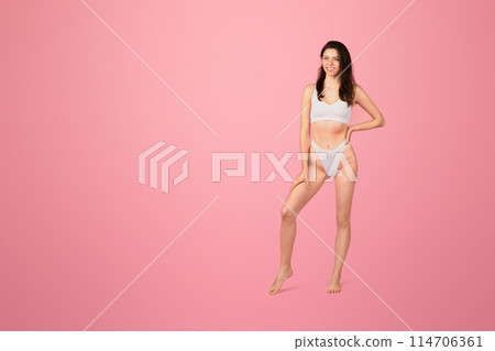 A radiant happy young woman with long brown hair poses confidently in a white two-piece swimsuit against a vibrant pink background, exuding fitness and health, full length 114706361