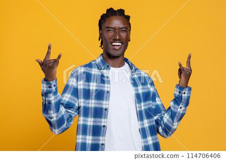 Energetic young black man showing rock and roll sign with hands and laughing, handsome african american guy wearing checkered shirt posing against bright yellow background, radiating fun Energetic young black man showing rock and roll sign with hands and laughing, handsome african american guy wearing checkered shirt posing against bright yellow background, radiating fun 114706406
