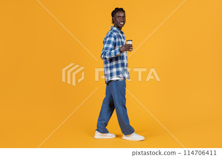 Relaxed young black man walking with takeaway coffee cup in hand, handsome african american guy dressed in plaid shirt and denim jeans posing on bright yellow background, copy space Relaxed young black man walking with takeaway coffee cup in hand, handsome african american guy dressed in plaid shirt and denim jeans posing on bright yellow background, copy space 114706411