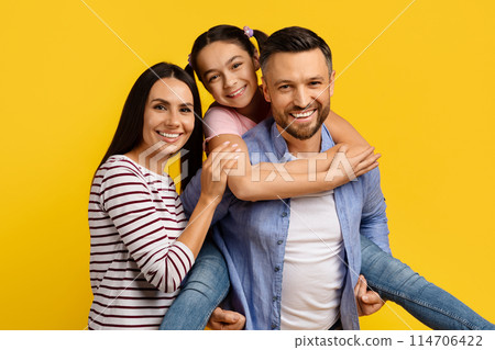 Loving family with daughter on father's back enjoying fun moment, hugging and smiling together, parents and cute teen female child posing together against vivid yellow studio background Loving family with daughter on father's back enjoying fun moment, hugging and smiling together, parents and cute teen female child posing together against vivid yellow studio background 114706422