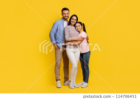 Happy caucasian family with teen daughter cuddling and smiling at camera, cheerful millennial parents and female kid dressed in casual clothes standing against bright yellow background, copy space 114706451