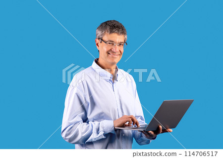 Smiling senior man in shirt confidently browsing online on his laptop, showcasing modern technology posing in blue studio setting, promoting e-learning or freelance opportunities 114706517