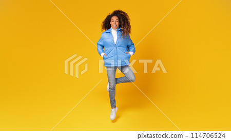 Stylish young black woman in blue winter jacket takes an energetic jump, set against vibrant yellow background, full length, panorama. Captures a holiday mood 114706524