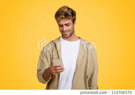 Stylish young European man is engrossed in texting on his cellphone, holding phone and looking at screen, standing posing against yellow studio background 114706570