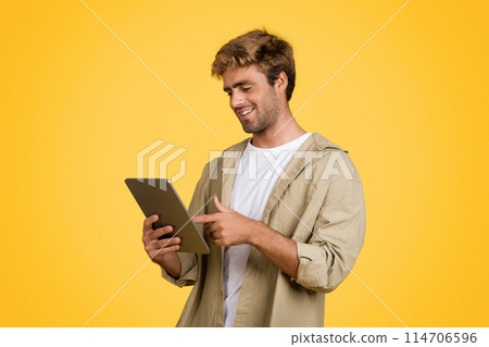 Stylish European man beams with contentment as he uses digital tablet, surfing the internet. He confidently stands against a striking yellow studio backdrop 114706596
