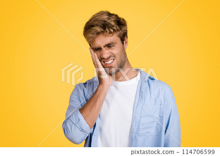Young European man showcases evident discomfort, touching his cheek due to toothache, clear depiction of dental pain on bright yellow studio backdrop 114706599