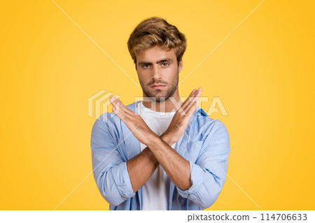 Serious young European man clearly gesturing no or refusal with decisive hand movement and facial expression, stands against vibrant yellow studio backdrop Serious young European man clearly gesturing no or refusal with decisive hand movement and facial expression, stands against vibrant yellow studio backdrop 114706633