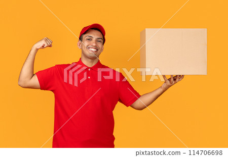 Power of fast delivery. Smiling mexican courier man in red uniform holds parcel carton box and showing biceps muscles on arm posing against yellow studio backdrop. Shipment offer 114706698