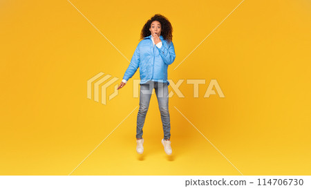 Surprised young black lady in winter jacket jumping, hand covering open mouth, against vivid yellow background, exuding startled holiday mood Surprised young black lady in winter jacket jumping, hand covering open mouth, against vivid yellow background, exuding startled holiday mood 114706730