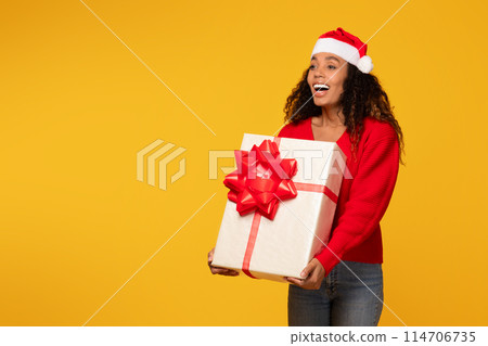 Radiant young black woman in Santa hat, proudly holding big wrapped Christmas gift, set against bright yellow background, free space Radiant young black woman in Santa hat, proudly holding big wrapped Christmas gift, set against bright yellow background, free space 114706735