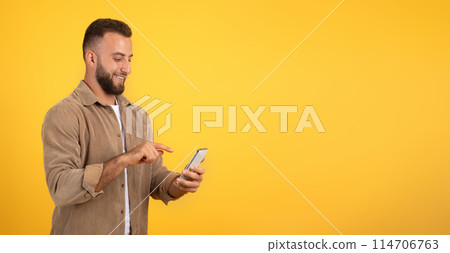 Positive european millennial guy with beard chatting on mobile phone, using app for blog, social networks, isolated on orange studio background, panorama. Work remotely, ad and offer 114706763