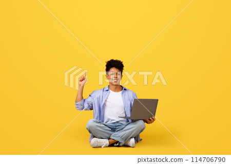 Stylish black student in studio setting uses laptop and points upward at empty space above him on vivid yellow background, symbolizing innovation and ideas 114706790