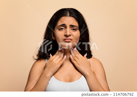 Portrait of unhappy sad sick indian woman suffering from sore throat, ill young eastern lady touching neck with hands, feeling ache, standing isolated over beige studio background, copy space 114706792